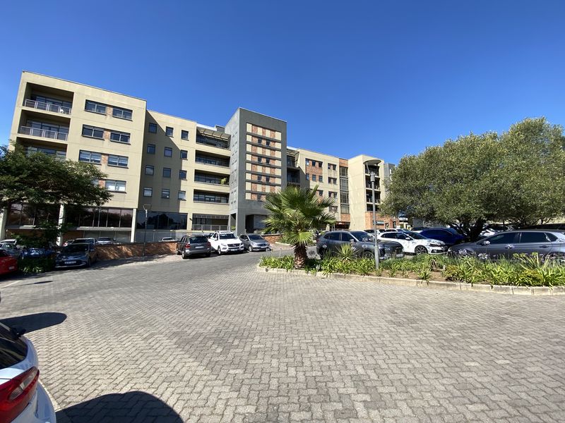 A wide-angle view of a modern multi-story office building featuring a paved parking area and landscaped garden beds.