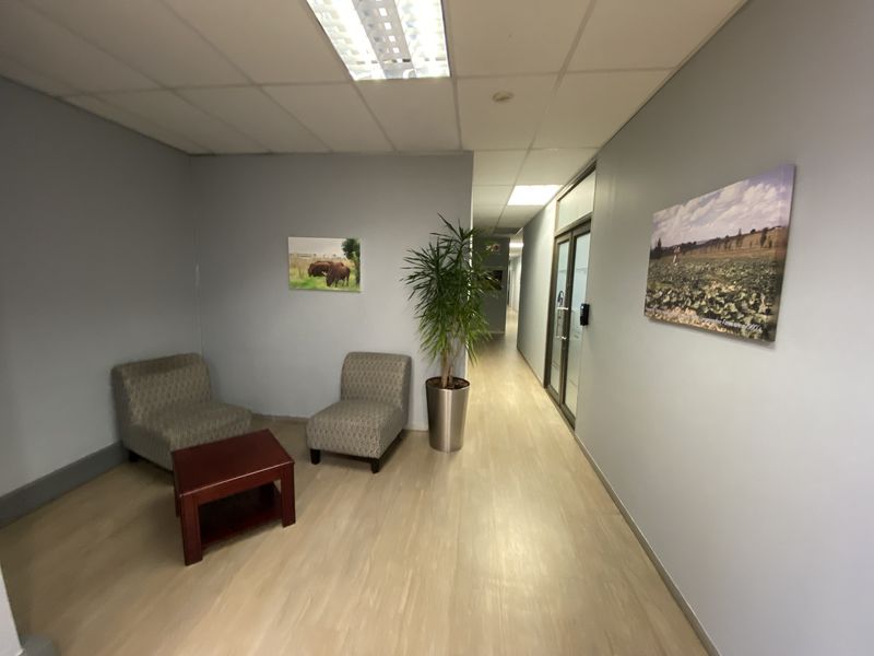 A functional office hallway featuring laminate flooring, suspended ceiling tiles with integrated lighting, and a glass-paneled door leading to adjacent office spaces.