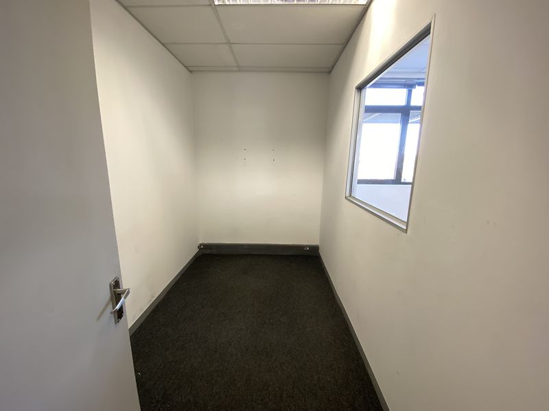 A compact, empty office space featuring a suspended ceiling, carpeted flooring, and an internal window providing natural light from the adjacent area.