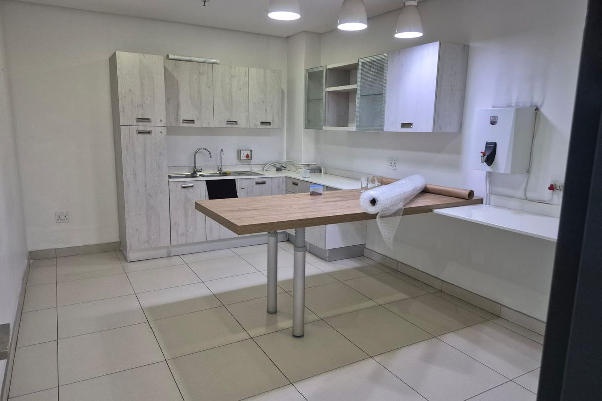 A functional kitchenette featuring light wood-grain cabinetry, a stainless steel sink, tiled flooring, and integrated lighting, suitable for office or industrial utility use.