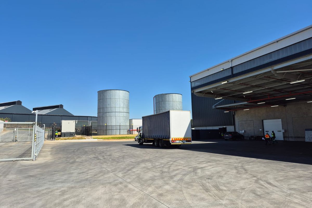Industrial facility exterior featuring a large concrete yard, metal storage silos, and a covered loading bay area with high-clearance access.
