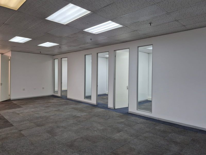 An open-plan office space featuring carpet tiles, a suspended ceiling with recessed lighting, and multiple glass-partitioned private office rooms.