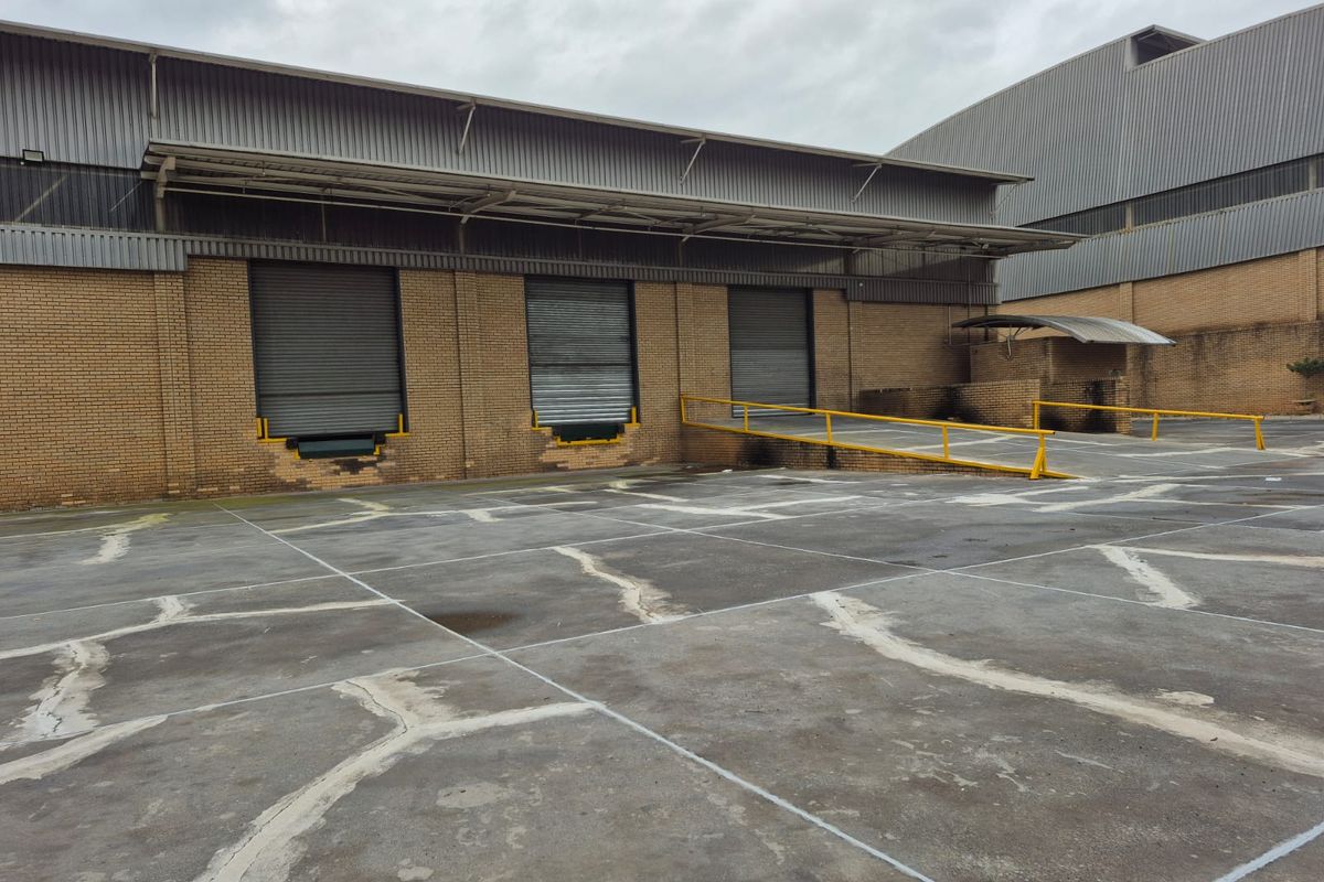 Industrial rear yard featuring multiple roller shutter loading bays, a concrete hardstand area, and protective steel safety railings.