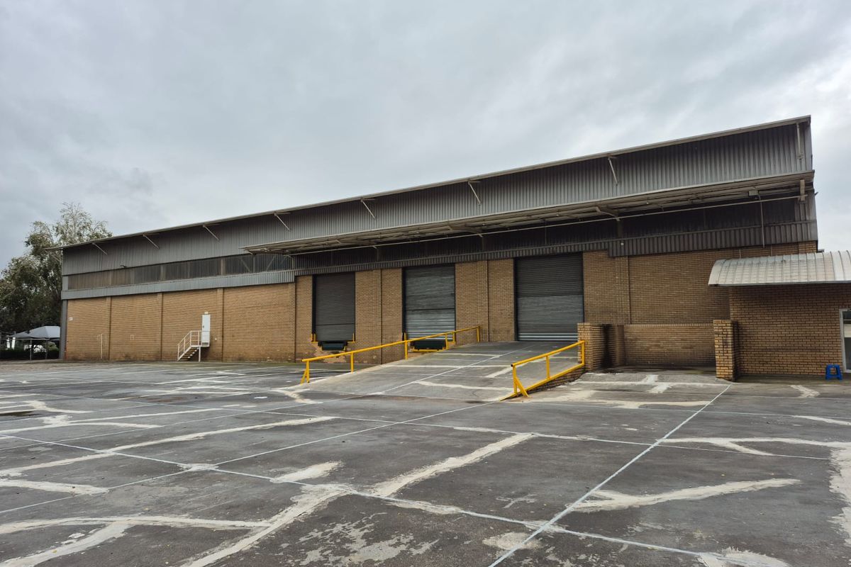 Industrial warehouse exterior featuring multiple roller shutter doors, a concrete loading ramp with safety railings, and a large asphalt yard area.