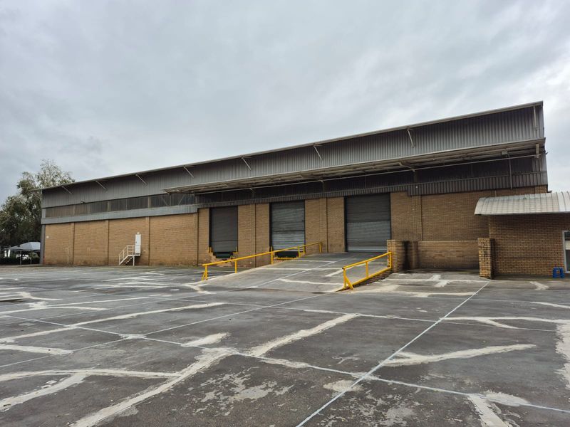 Industrial warehouse exterior featuring multiple roller shutter doors, a concrete loading ramp with safety railings, and a large asphalt yard area.