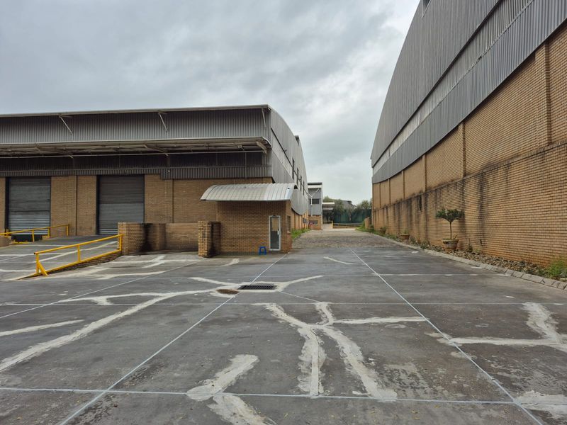 Industrial exterior yard area featuring concrete paving, loading docks with roller shutter doors, and brick warehouse structures.