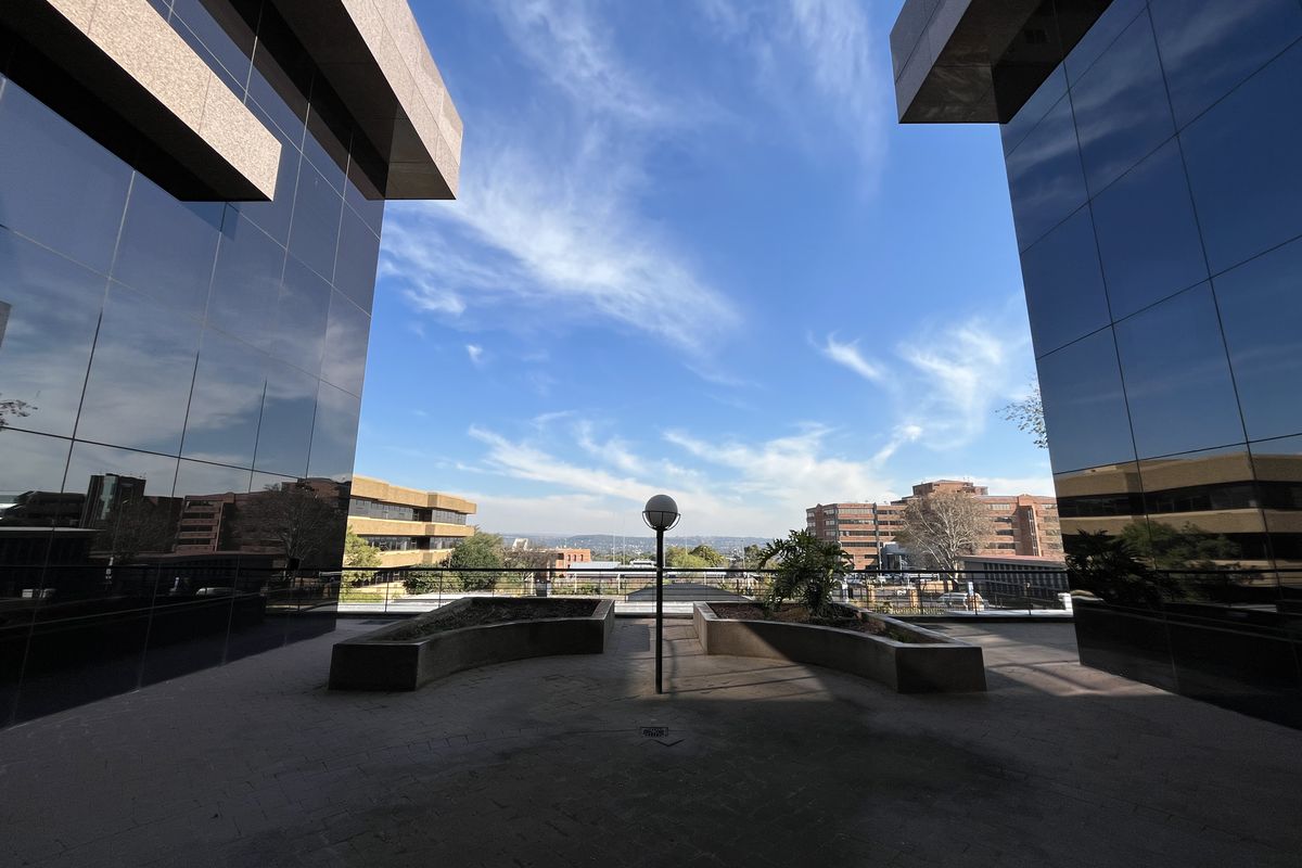 A modern office complex courtyard featuring reflective glass facades, paved flooring, and built-in concrete planters with a clear view of the surrounding urban landscape.