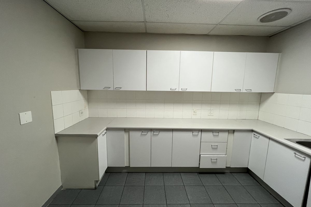 A functional office kitchenette featuring white cabinetry, laminate countertops, tiled backsplash, and a drop ceiling with integrated lighting.