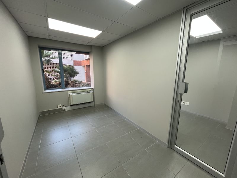 A clean, empty office space featuring tiled flooring, a suspended ceiling with recessed lighting, an HVAC unit, and an aluminum-framed glass entrance door.