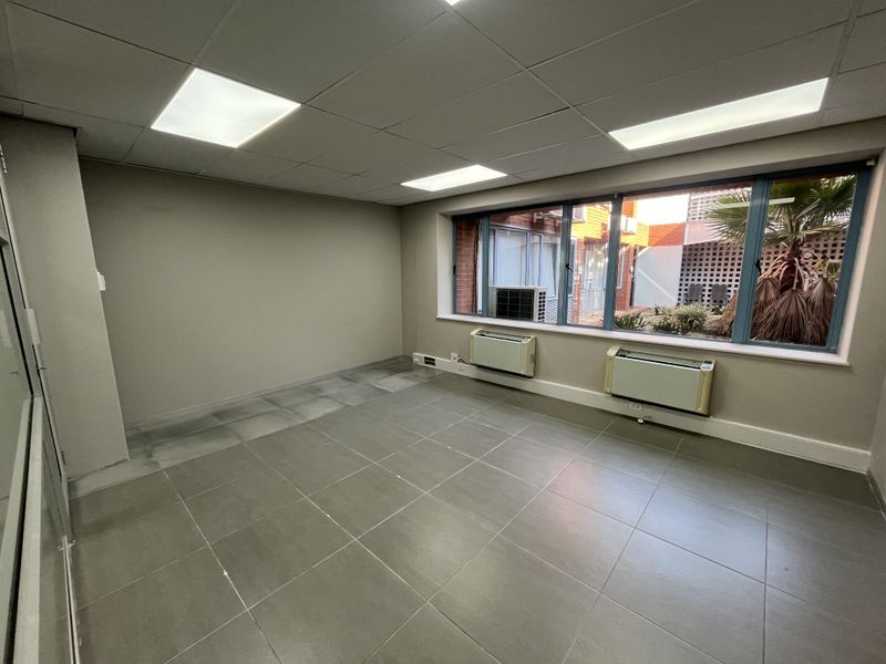 A vacant office space featuring tiled flooring, a suspended ceiling with recessed lighting, and large windows with wall-mounted HVAC units.