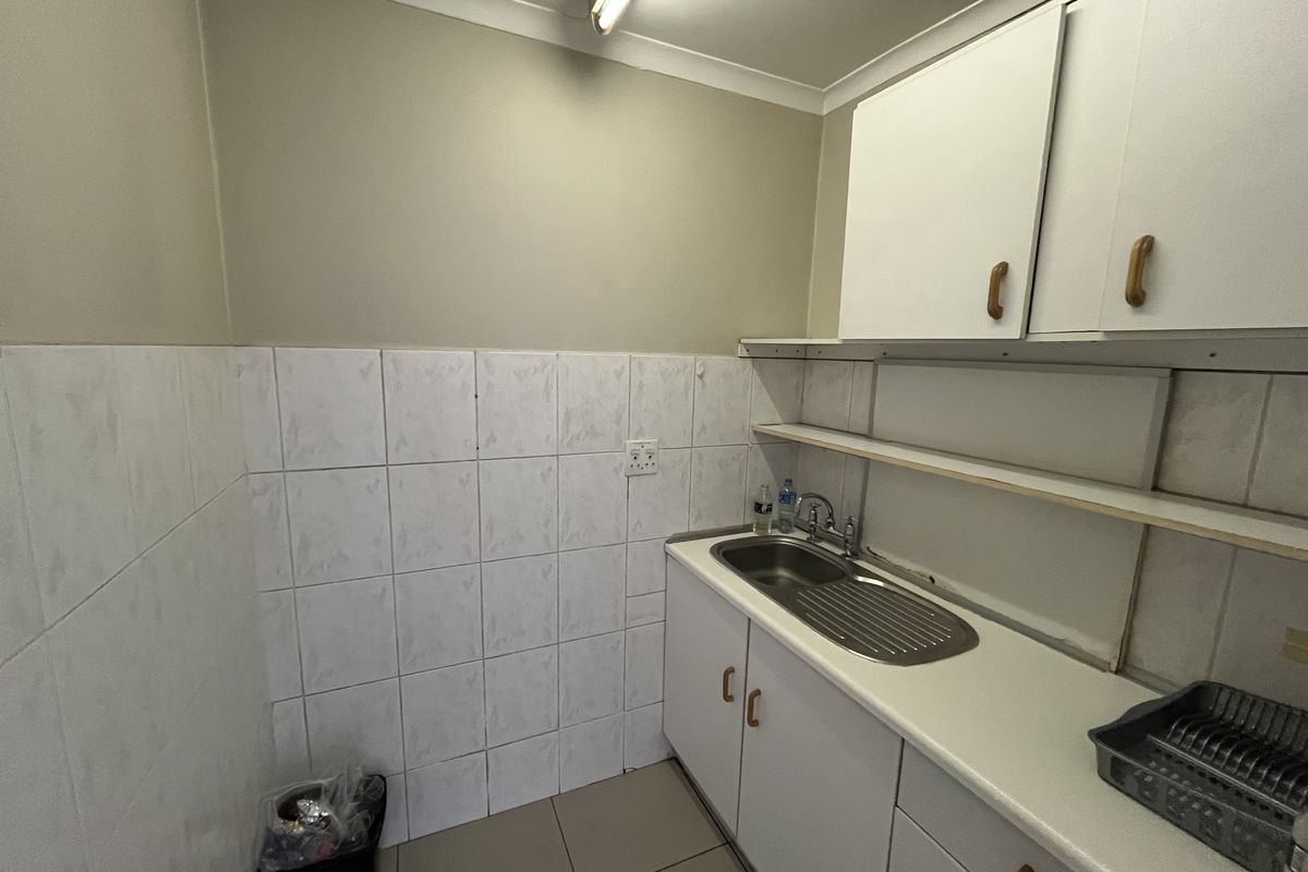 A functional office kitchenette featuring white cabinetry, a stainless steel sink, tiled splashbacks, and open shelving for storage.