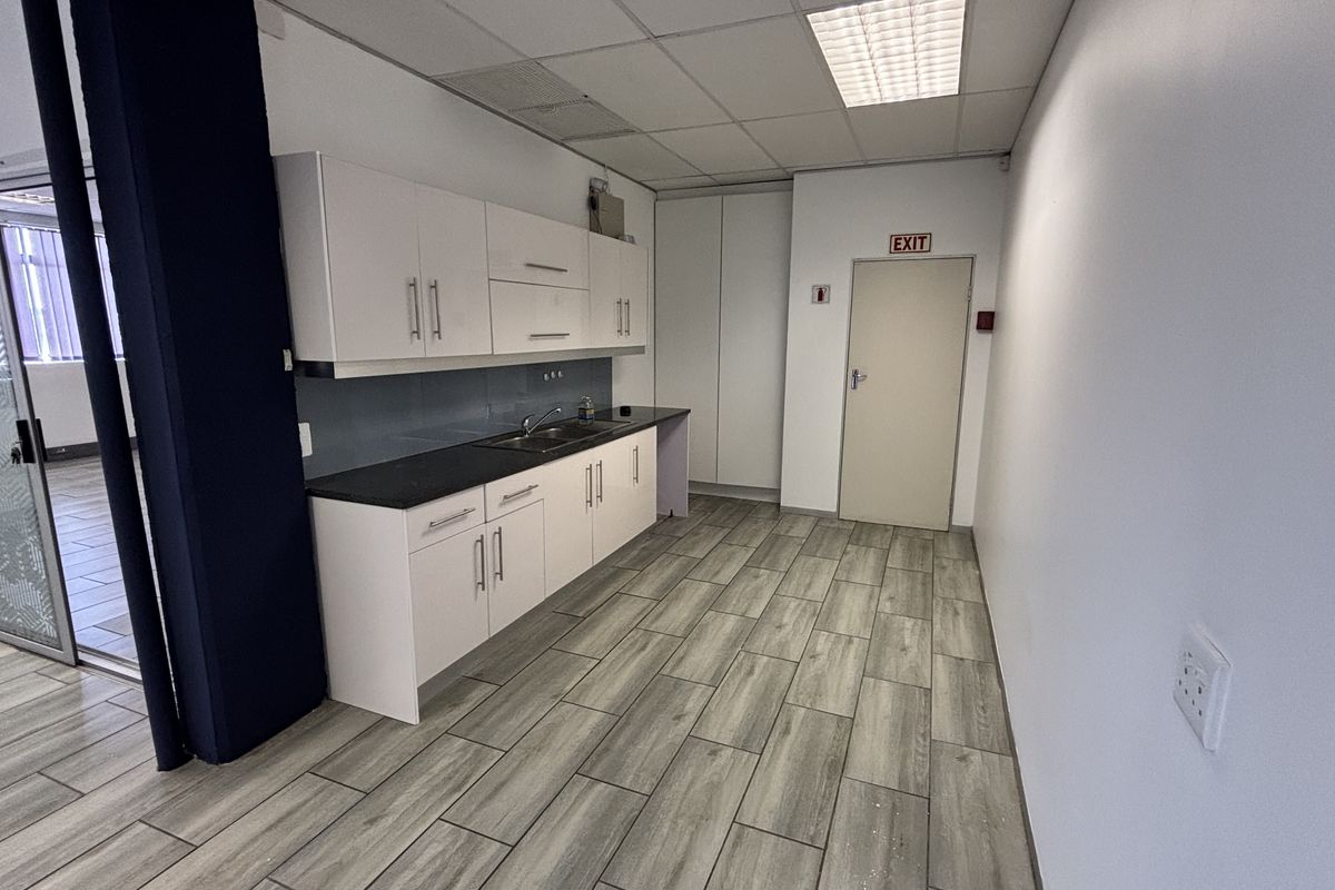 A clean, functional office kitchenette featuring white cabinetry, dark countertops, stainless steel sink, and durable wood-look tile flooring.