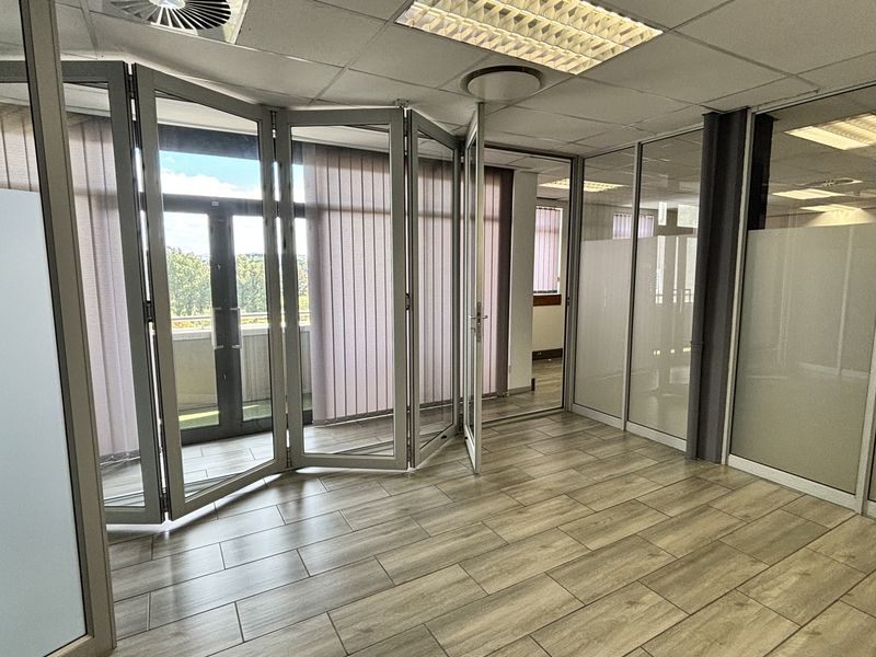 Modern office space featuring wood-look tile flooring, suspended ceiling, and extensive aluminium-framed glass partitioning with folding doors leading to an outdoor area.