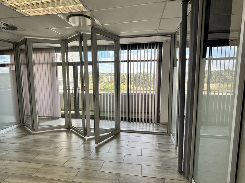 Modern office space featuring glass partition walls, folding doors leading to a balcony, tiled flooring, and recessed lighting in a suspended ceiling.