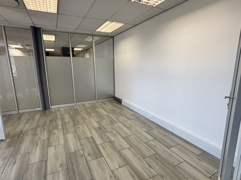 A clean, empty office space featuring modern wood-look tile flooring, glass partition walls, and a suspended ceiling with recessed lighting.