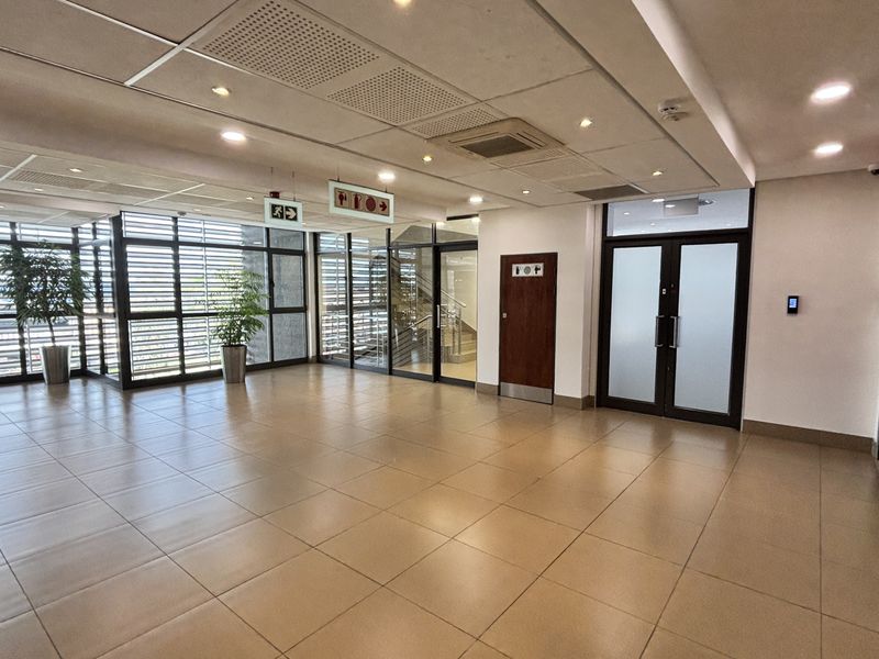 A spacious, modern office lobby or reception area featuring tiled flooring, suspended ceilings with recessed lighting, and glass partition walls.