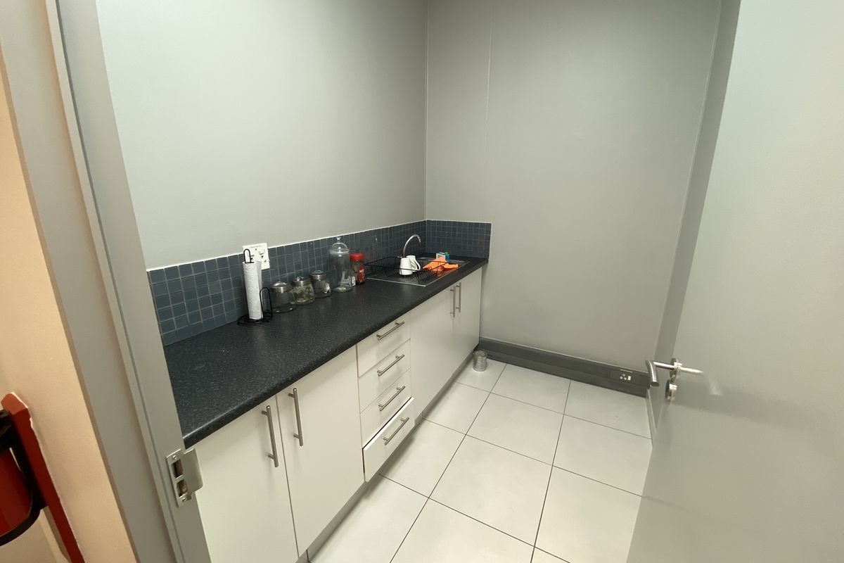 A compact office kitchenette featuring white cabinetry, dark laminate countertops, a stainless steel sink, and tiled splashback.