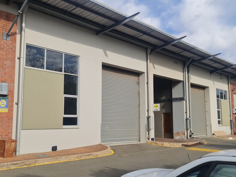 Front elevation of an industrial warehouse complex featuring multiple roller shutter doors, brick and rendered facade, and a functional paved driveway area.