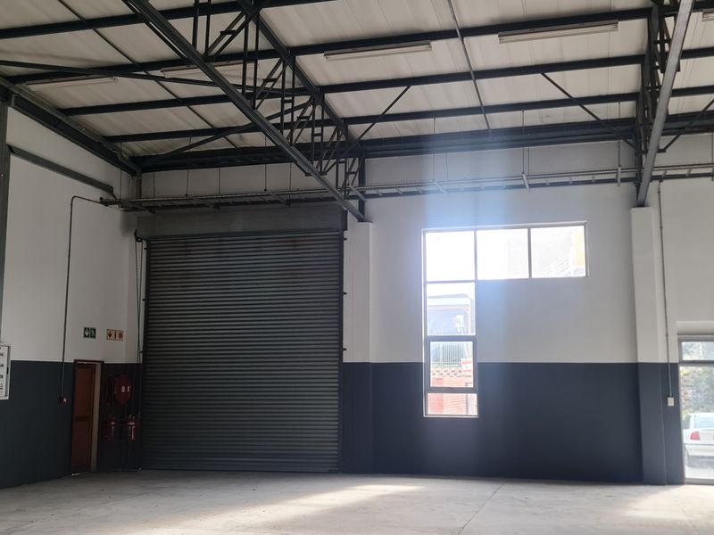Spacious industrial warehouse unit featuring high ceilings with exposed steel trusses, a large roller shutter door, and concrete flooring.