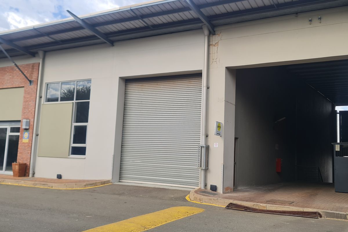 Industrial property exterior featuring a roller shutter door, glass entrance, and paved access area under a metal roof structure.