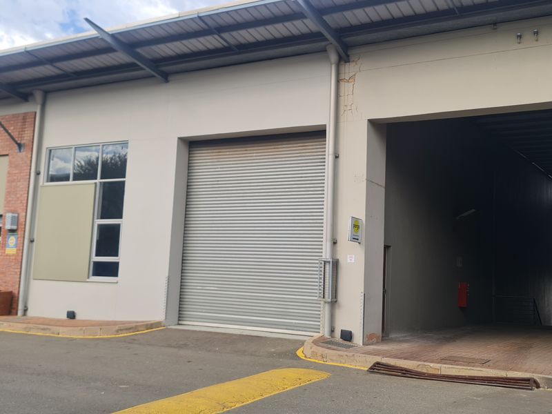 Industrial property exterior featuring a roller shutter door, glass entrance, and paved access area under a metal roof structure.