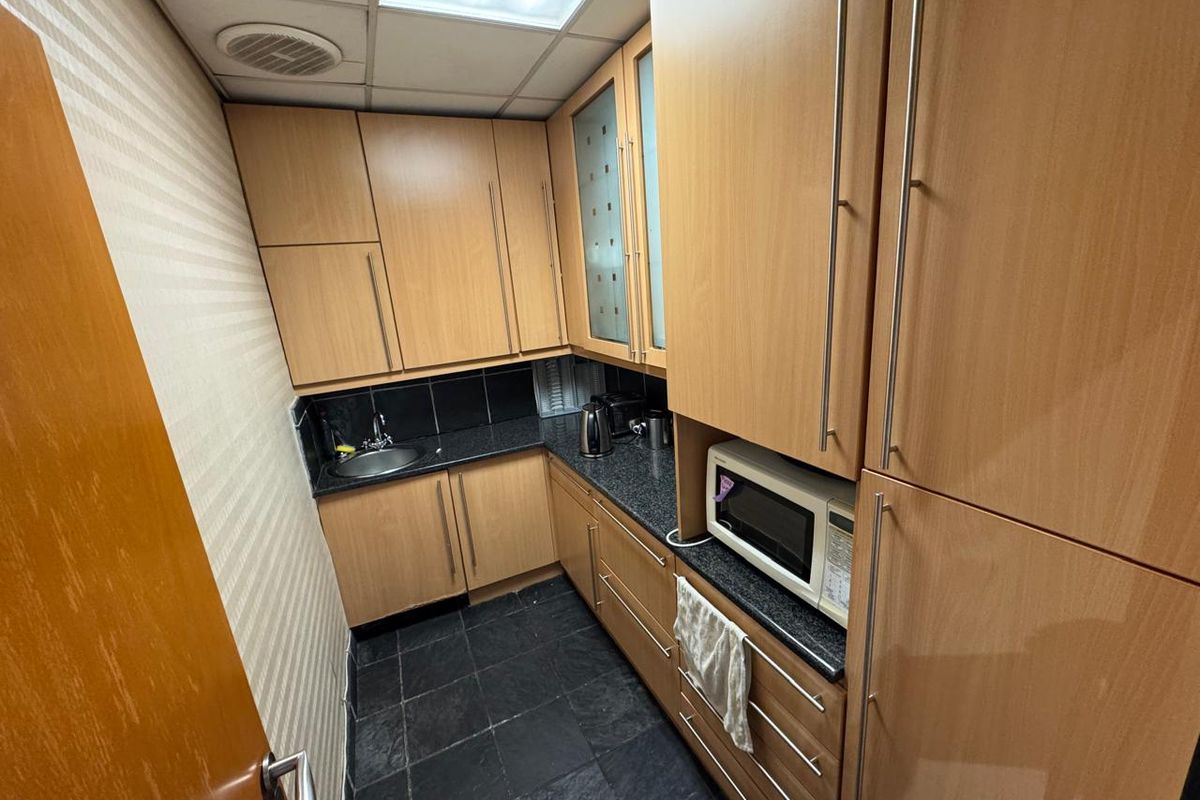 A compact office kitchenette featuring wood-finish cabinetry, dark stone-look countertops, a stainless steel sink, and durable tiled flooring.