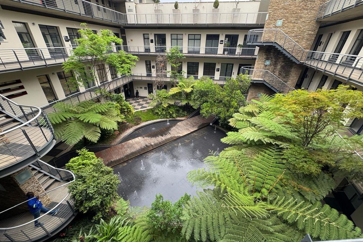 A high-angle view of a modern office complex courtyard featuring a central water feature, wooden footbridge, lush landscaping, and multi-level access balconies.