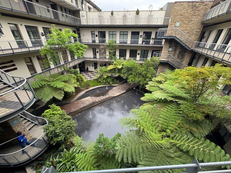 A high-angle view of a modern office complex courtyard featuring a central water feature, wooden footbridge, lush landscaping, and multi-level access balconies.