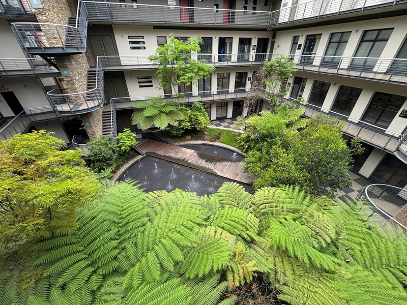 A well-maintained central courtyard featuring a water feature, wooden footbridge, and lush landscaping surrounded by multi-level office building walkways.