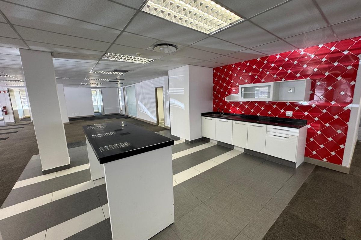 A functional office kitchenette featuring white cabinetry, a black countertop, and a distinctive red tiled backsplash, integrated into an open-plan workspace.