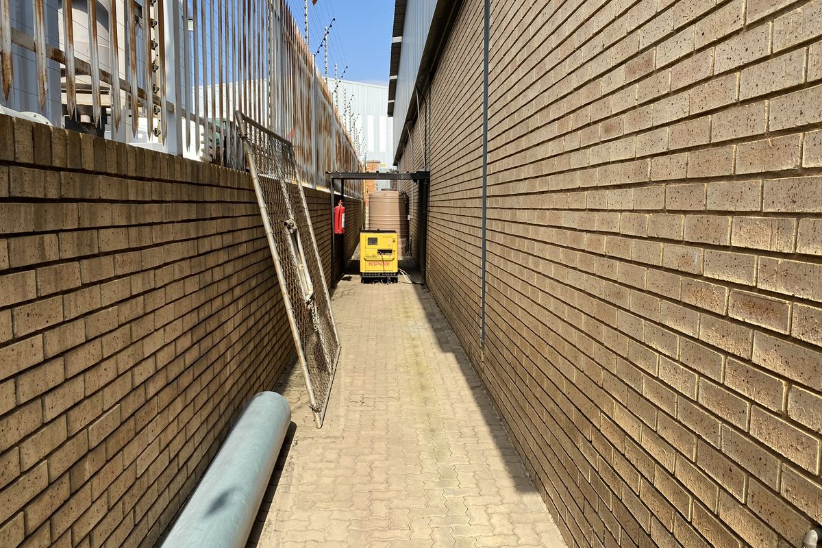 A narrow exterior side passage featuring brick walls, paved flooring, and security fencing, providing access to utility infrastructure.