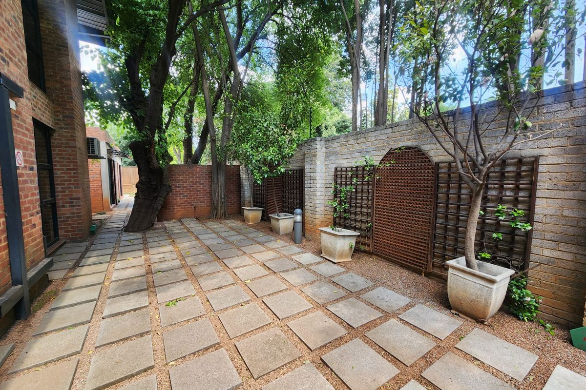 A paved outdoor courtyard area featuring brick boundary walls, wooden trellis panels, and mature trees, providing a private and tranquil exterior space.
