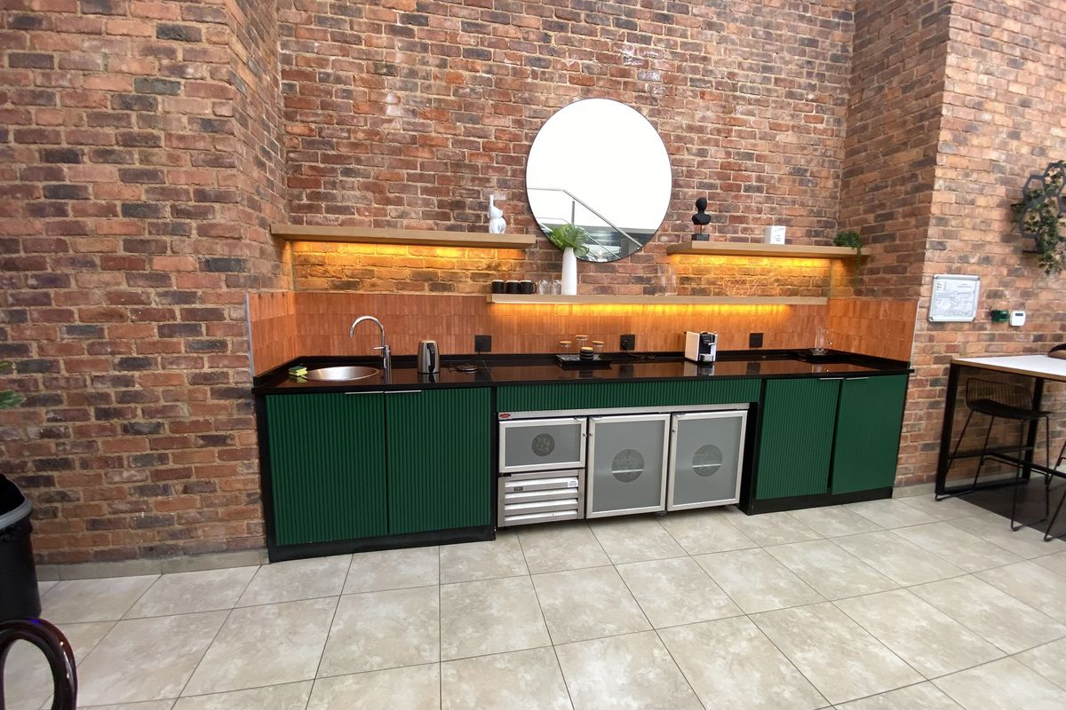 A modern office kitchenette featuring green fluted cabinetry, a black countertop, integrated under-counter refrigeration, and warm LED accent lighting against an exposed brick wall.
