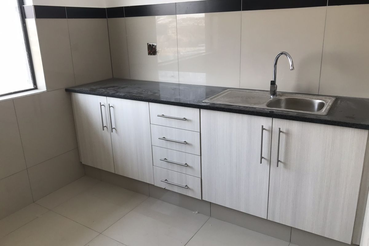 A clean, modern kitchen kitchenette featuring light wood-grain cabinetry, dark granite countertops, a stainless steel sink, and neutral wall tiling with a decorative border.