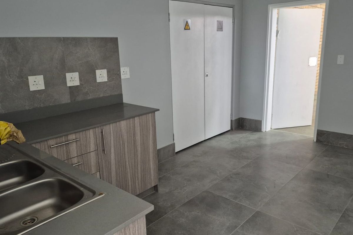 A clean, modern industrial kitchen area featuring stone-look floor tiles, laminate cabinetry, a double stainless steel sink, and integrated electrical distribution board access.