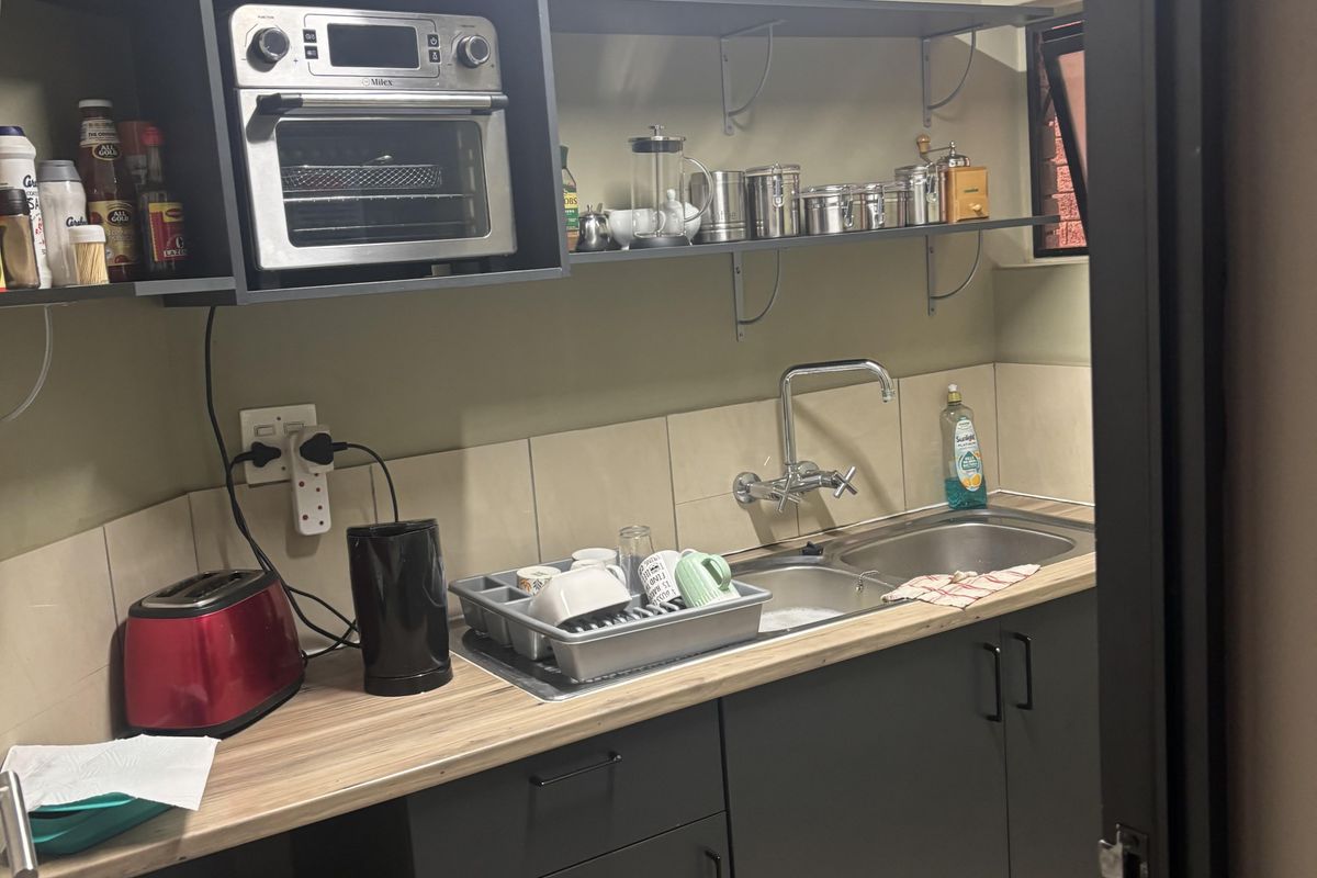 Compact office kitchenette featuring dark cabinetry, a stainless steel double sink, tiled backsplash, and open shelving for storage.