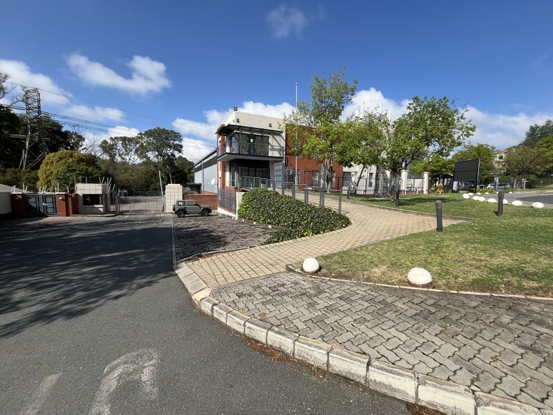 Front exterior view of an industrial property featuring a paved driveway, secure gated entrance, landscaped garden areas, and a multi-story building with balcony.