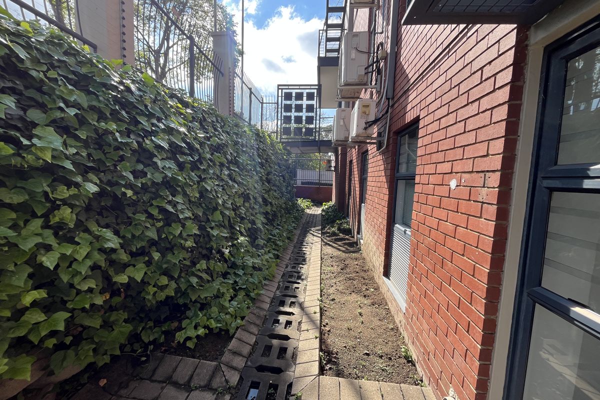 A narrow exterior side passage featuring red brick walling, paved drainage channels, and mounted air conditioning units, secured by perimeter electric fencing.