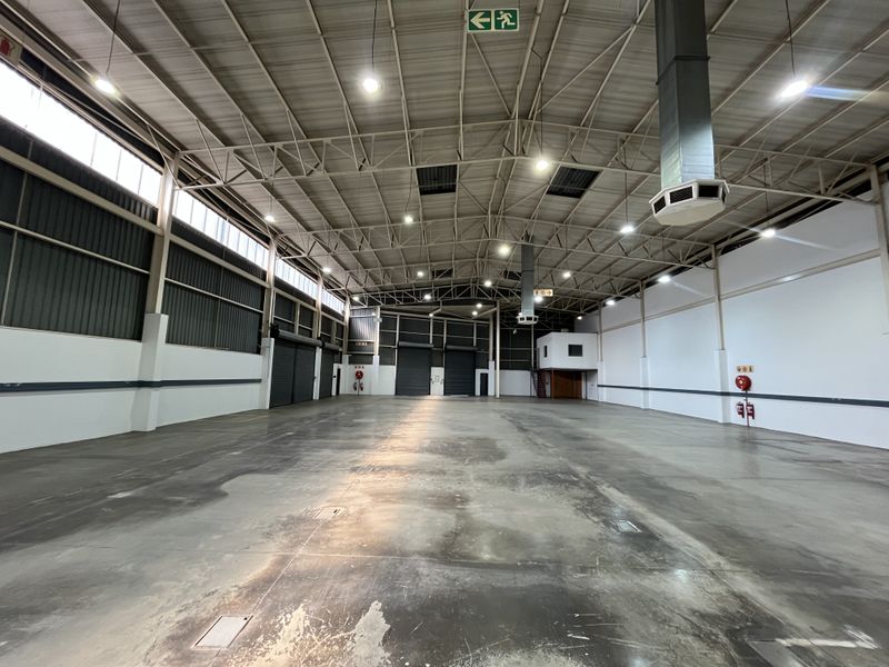 Spacious industrial warehouse featuring high ceilings, exposed steel trusses, roller shutter doors, and durable concrete flooring, suitable for various storage or manufacturing operations.