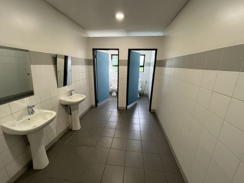 A clean, functional industrial-style communal bathroom featuring dual pedestal wash basins, tiled walls and flooring, and two separate toilet cubicles.
