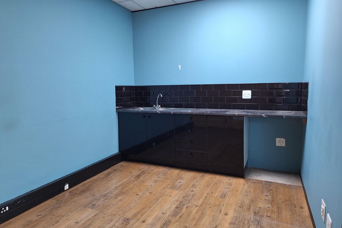 A small office kitchenette featuring dark cabinetry, a black tiled backsplash, a stainless steel sink, and wood-look flooring.