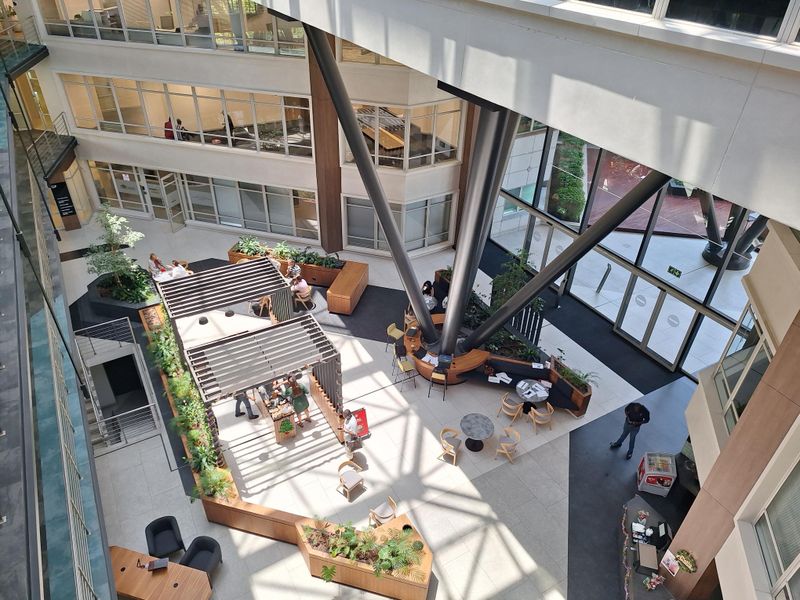 A high-angle view of a modern, multi-level office atrium featuring large structural support columns, glass facades, and integrated indoor landscaping.