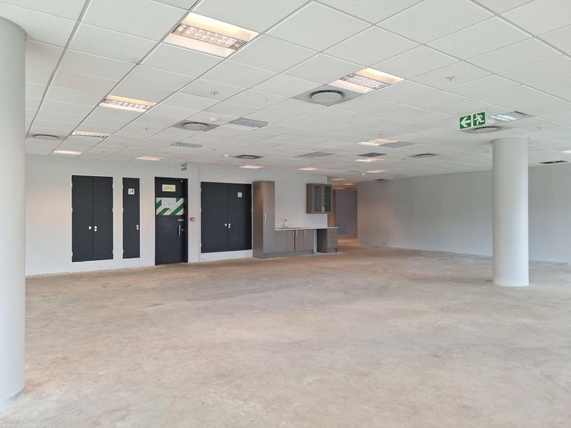 A spacious, open-plan office area featuring a suspended ceiling with recessed lighting, integrated kitchenette, and structural support columns.