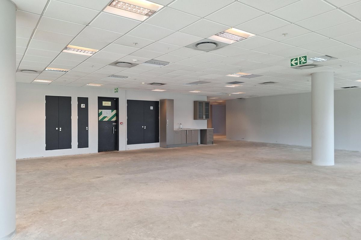 A spacious, open-plan office area featuring a suspended ceiling with recessed lighting, integrated kitchenette, and structural support columns.