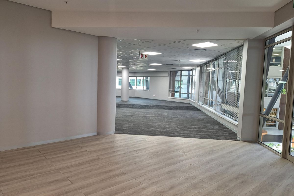 Spacious open-plan office area featuring a mix of wood-look laminate flooring and carpet, large windows with blinds, and a suspended ceiling grid.