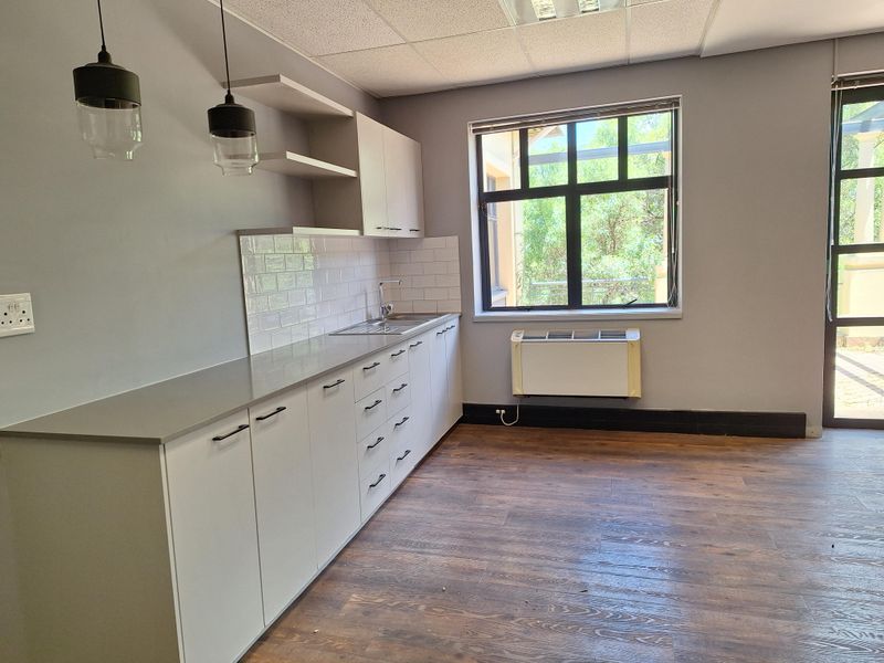 A clean, modern office kitchenette featuring white cabinetry, subway tile backsplash, stainless steel sink, and wood-look flooring with natural light.