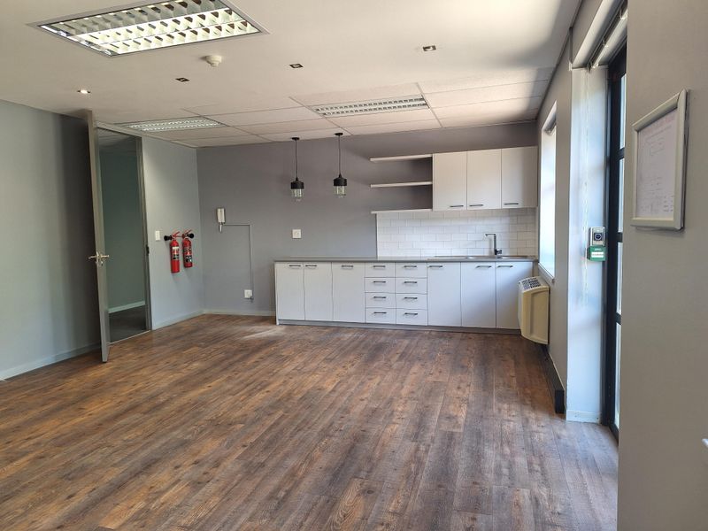 A bright, open-plan office space featuring modern laminate flooring, a functional kitchenette with built-in cabinetry, and professional recessed lighting.