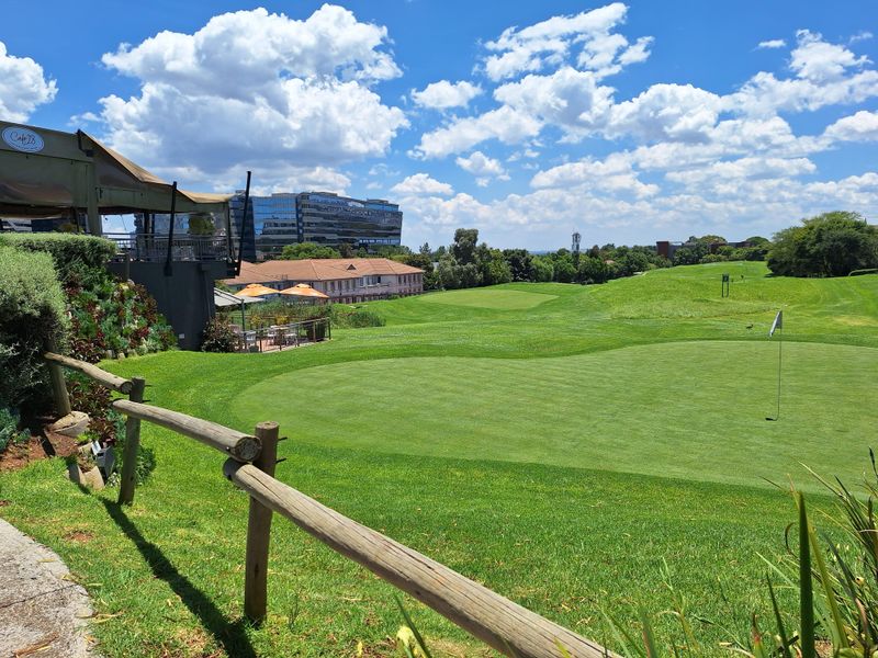 A scenic view overlooking a well-maintained golf course with a timber fence in the foreground and commercial buildings in the distance.