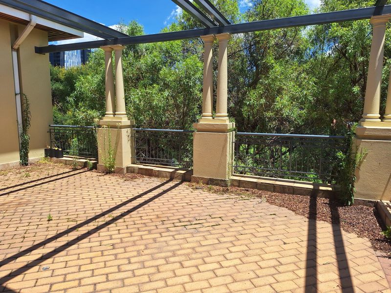 A spacious outdoor patio area featuring brick paving, a metal pergola structure, and decorative columns with metal railings overlooking lush greenery.