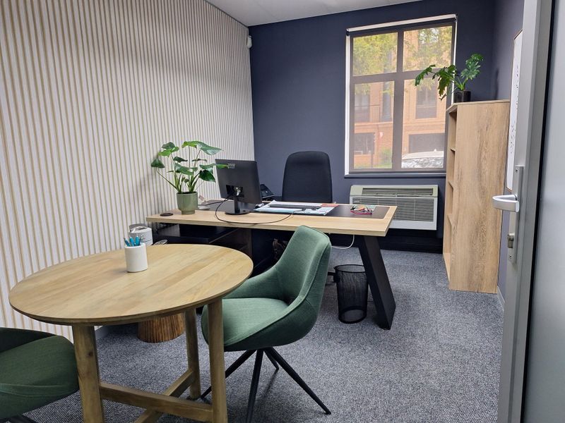 A professional office space featuring a window with natural light, carpeted flooring, and a wall-mounted HVAC unit.